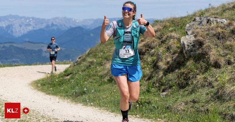 Eine Frau in blauer und grauer Sportbekleidung läuft und gibt einen Daumen hoch. Sie trägt ein Startnummernband mit der Nummer 13. Hinter ihr läuft ein Mann auf einem Bergpfad.