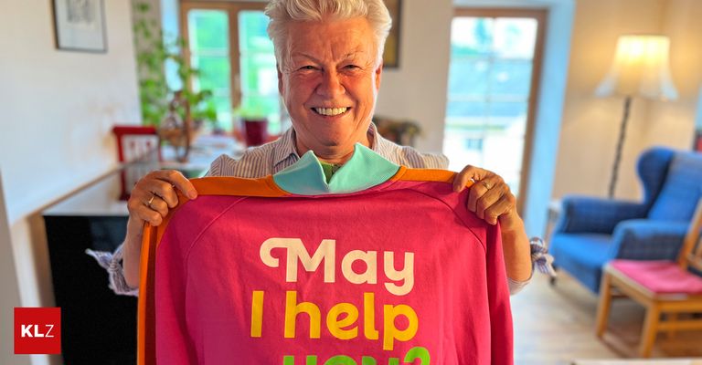 An elderly person holding a pink shirt that says 'May I help you?' smiling in a room with natural light and indoor plants.
