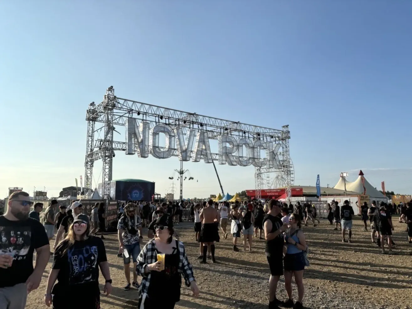 Eine große Menschenmenge versammelt sich beim Nova Rock Festival mit einer Metallbogenstruktur im Hintergrund. Die Leute gehen und stehen herum, einige tragen Sonnenbrillen und Hüte.