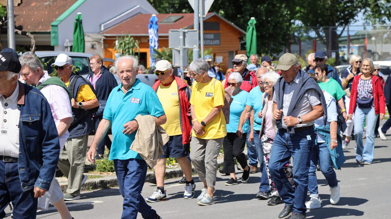 Eine Gruppe älterer Menschen geht die Straße entlang, einige tragen Hüte, Brillen und Turnschuhe. Sie sind von Gebäuden und Grün umgeben, und eine Person hält einen Mantel.