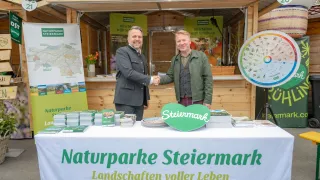 Zwei Männer schütteln sich die Hände vor einem Stand. Ein Mann trägt einen Anzug, der andere einen grünen Pullover. Ein Tisch mit Büchern und einem Schild mit der Aufschrift 'Naturparke Steiermark' steht im Vordergrund.