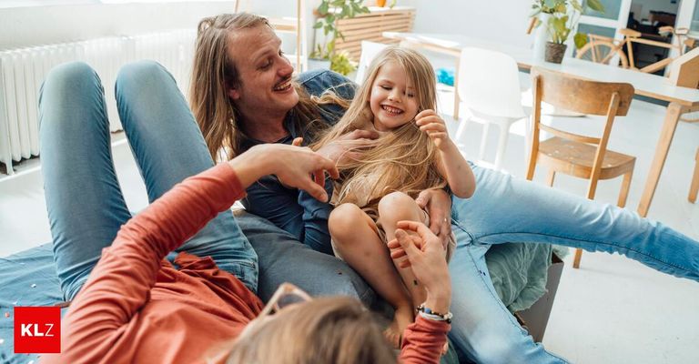 Eine Familie sitzt auf einer Couch, ein Mann kitzelt ein junges Mädchen, das lacht, während ein anderes Kind zusieht.