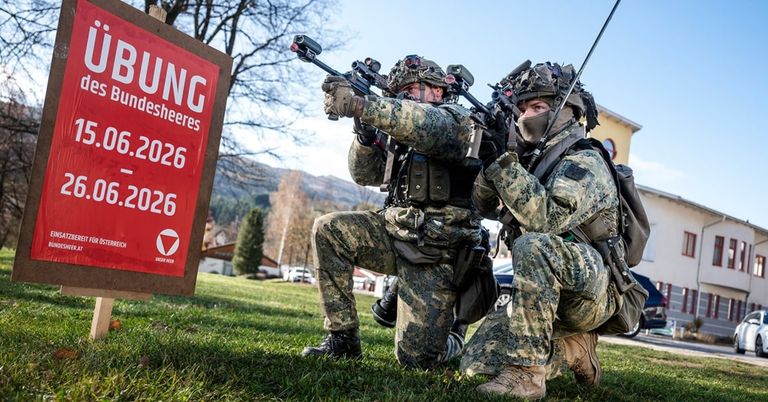 Zwei Soldaten kauen auf dem Gras und halten Gewehre. Einer zielt mit seiner Waffe auf die Kamera und der andere schaut nach vorne. Ein rotes Schild mit weißem Text und einem Logo steht hinter ihnen.