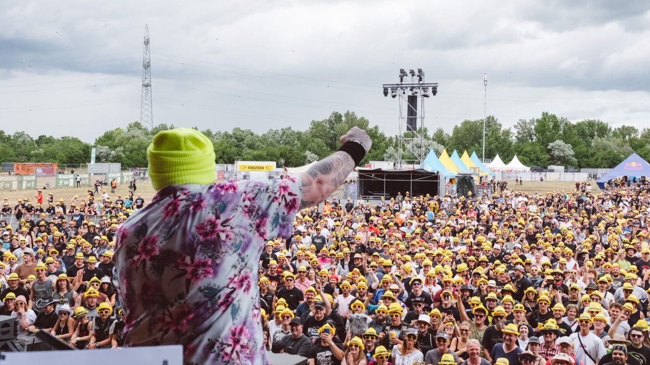 Ein Mann in einem Blumenhemd und gelbem Hut hebt seinen Arm bei einem Open-Air-Festival. Die Menge trägt gelbe Hüte und im Hintergrund befindet sich eine Bühne mit einer Lichtsäule.
