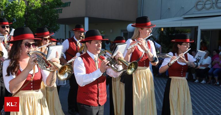 Eine Blaskapelle in traditioneller Kleidung spielt im Freien. Eine Frau spielt Flöte, ein Mann Trompete. Hinter ihnen stehen Zuschauer.