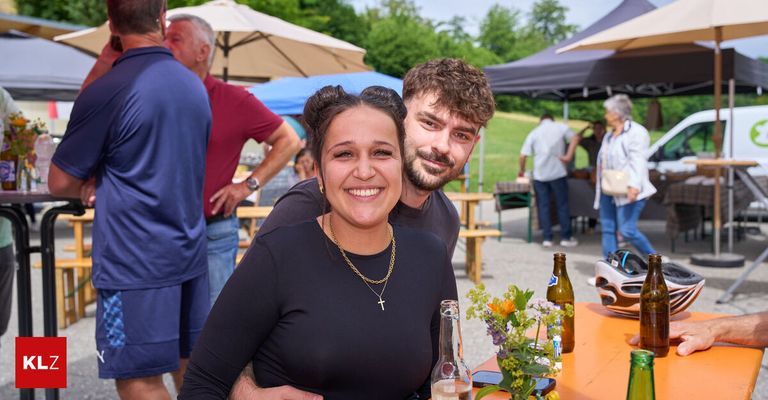 Ein lächelndes Paar steht neben einem Tisch mit Blumen und Flaschen, bei einer Outdoor-Veranstaltung mit Zelten und Menschen im Hintergrund.