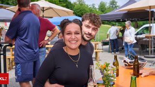 Ein lächelndes Paar steht neben einem Tisch mit Blumen und Flaschen, bei einer Outdoor-Veranstaltung mit Zelten und Menschen im Hintergrund.