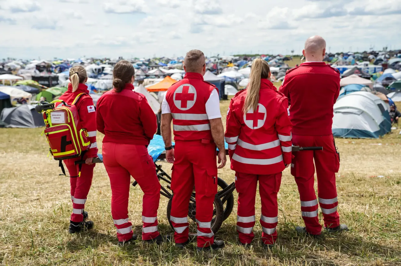 Eine Gruppe von Rettungskräften in roten Uniformen mit einem roten Kreuzsymbol auf dem Rücken steht auf einem Grasfeld. Sie blicken in die Ferne mit Zelten und einer Menge.