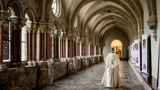 Ein Priester geht allein einen langen, gepflasterten Gang einer Kirche mit gewölbten Decken und Rundfenstern entlang.