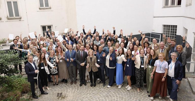 Eine Gruppe von Menschen, die für ein Foto in einer großen Versammlung posieren, wobei einige Daumen hochhalten und andere Papiere halten. Sie sind in formeller und lässiger Kleidung gekleidet.