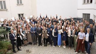 Eine Gruppe von Menschen, die für ein Foto in einer großen Versammlung posieren, wobei einige Daumen hochhalten und andere Papiere halten. Sie sind in formeller und lässiger Kleidung gekleidet.