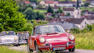 Ein roter Oldtimer fährt auf einer Straße umgeben von Grün, mit einem Dorf im Hintergrund.