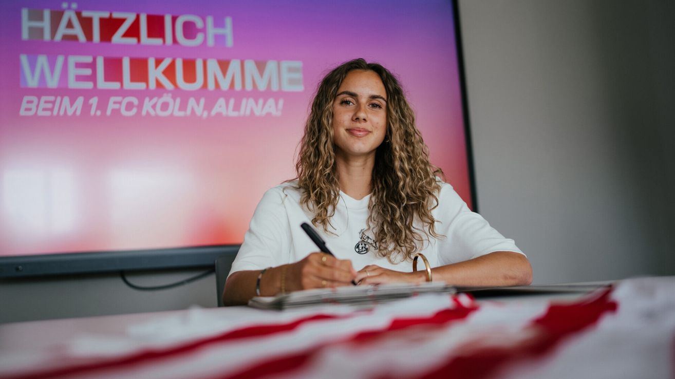 Eine Frau mit lockigem Haar sitzt an einem Tisch, lächelt und hält einen Stift. Sie signiert wahrscheinlich Autogramme. Hinter ihr zeigt eine große Leinwand pinken und roten Text.