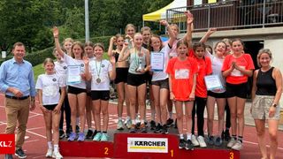 Eine Gruppe junger Mädchen in Sportuniformen steht auf einem Podest bei einem Leichtathletikevent, lächelt und hält Medaillen. Das Podest hat die Zahlen 1, 2 und 3 und das Wort 'Kärcher'. Sie stehen auf der Laufbahn, mit einem Zaun und Bäumen im Hintergrund.