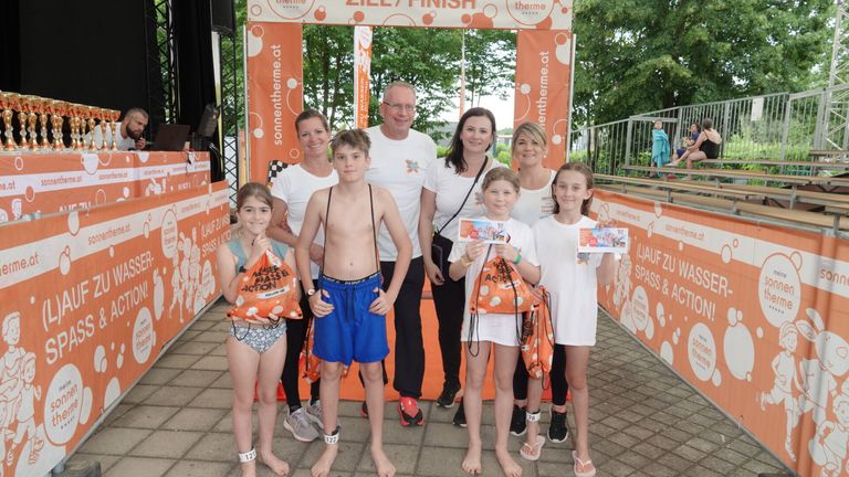 Eine Familie posiert für ein Bild bei einem Marathon-Event, alle in weißen T-Shirts und mit orangefarbenen Taschen. Dahinter stehen zwei Erwachsene, und ein Banner steht 'Ziel/Finish'.