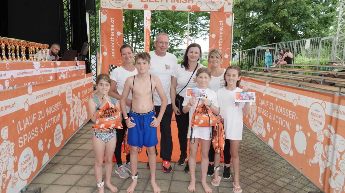 Eine Familie posiert für ein Bild bei einem Marathon-Event, alle in weißen T-Shirts und mit orangefarbenen Taschen. Dahinter stehen zwei Erwachsene, und ein Banner steht 'Ziel/Finish'.