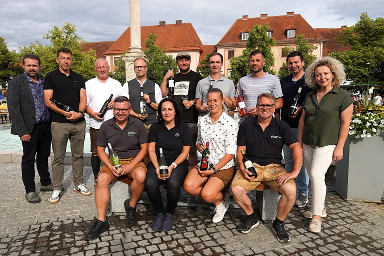 Eine Gruppe von Menschen posiert mit Weinflaschen auf einem historischen Stadtplatz vor Gebäuden im Hintergrund.