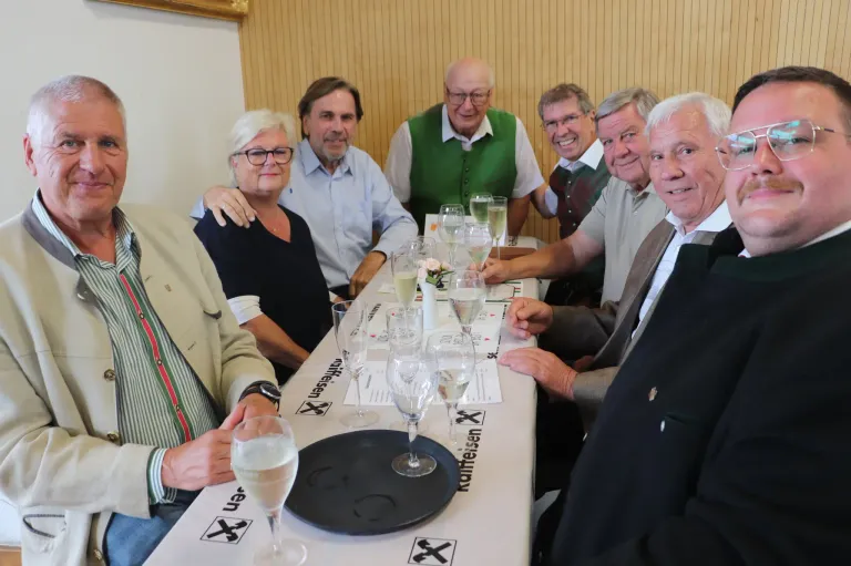 Eine Gruppe älterer Menschen sitzt an einem Tisch mit Weinläsern und lächelt. Sie halten einen schwarzen Teller mit einem Kreuzsymbol. Der Tisch hat eine weiße Tischdecke mit dem Namen der Veranstaltung.