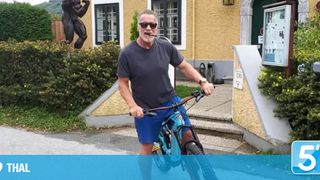 Ein älterer Mann in einem grauen T-Shirt und blauer Shorts fährt ein blaues Fahrrad vor einem Haus mit einer Statue eines Mannes, der Gewichte hebt.