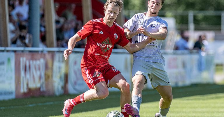 Zwei Fußballspieler konkurrieren auf einem Feld. Einer trägt ein rotes Trikot mit HOLZ STEF und der andere trägt ein weißes Trikot mit FCPS. Sie befinden sich dabei, sich gegenseitig in der Nähe des Balls zu tackeln.