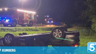 Ein umgestürztes Auto liegt auf der Seite in einem Grasbereich, mit Notfallfahrzeugen in der Nähe. Ein Feuerwehrwagen ist geparkt, und ein weiteres Notfallfahrzeug ist auf der Straße.