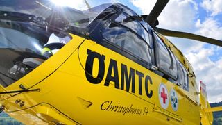 Ein gelber Hubschrauber, beschriftet mit 'DAMTC Christophorus 14', steht unter einem teilweise bewölkten Himmel, mit der Sonne, die darauf scheint.
