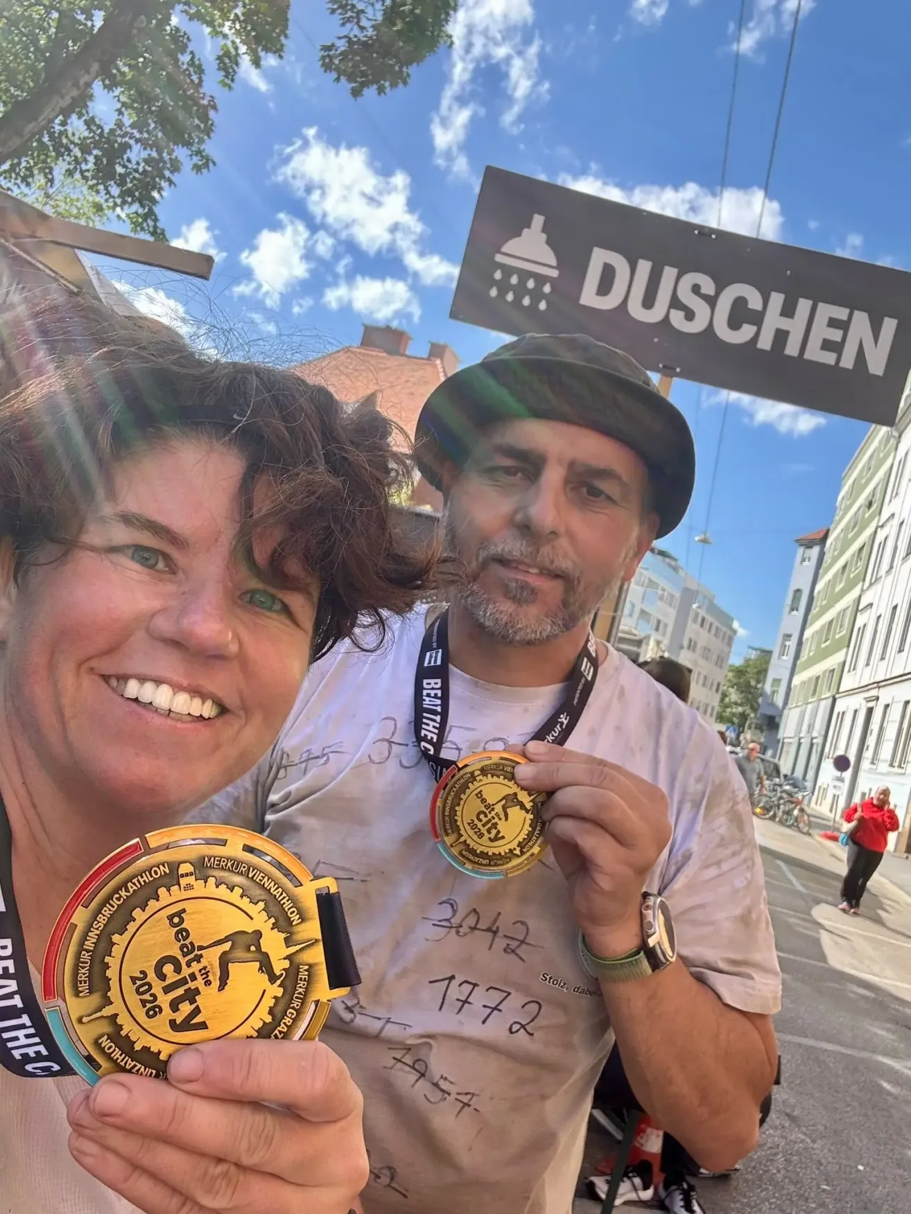 Ein Mann und eine Frau posieren für ein Foto mit Marathon-Medaillen. Ein Schild für Duschen steht hinter ihnen.