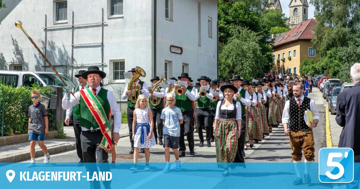 Ein traditionelles Musikorchester marschiert die Straße entlang, mit Musikern in grünen Westen, angeführt von einem Mann mit einer Schärpe. Kinder in bunten Kleidern gesellen sich zur Prozession. Gebäude säumen die Straße, mit Grün im Hintergrund.