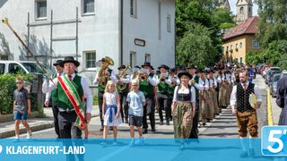 Ein traditionelles Musikorchester marschiert die Straße entlang, mit Musikern in grünen Westen, angeführt von einem Mann mit einer Schärpe. Kinder in bunten Kleidern gesellen sich zur Prozession. Gebäude säumen die Straße, mit Grün im Hintergrund.