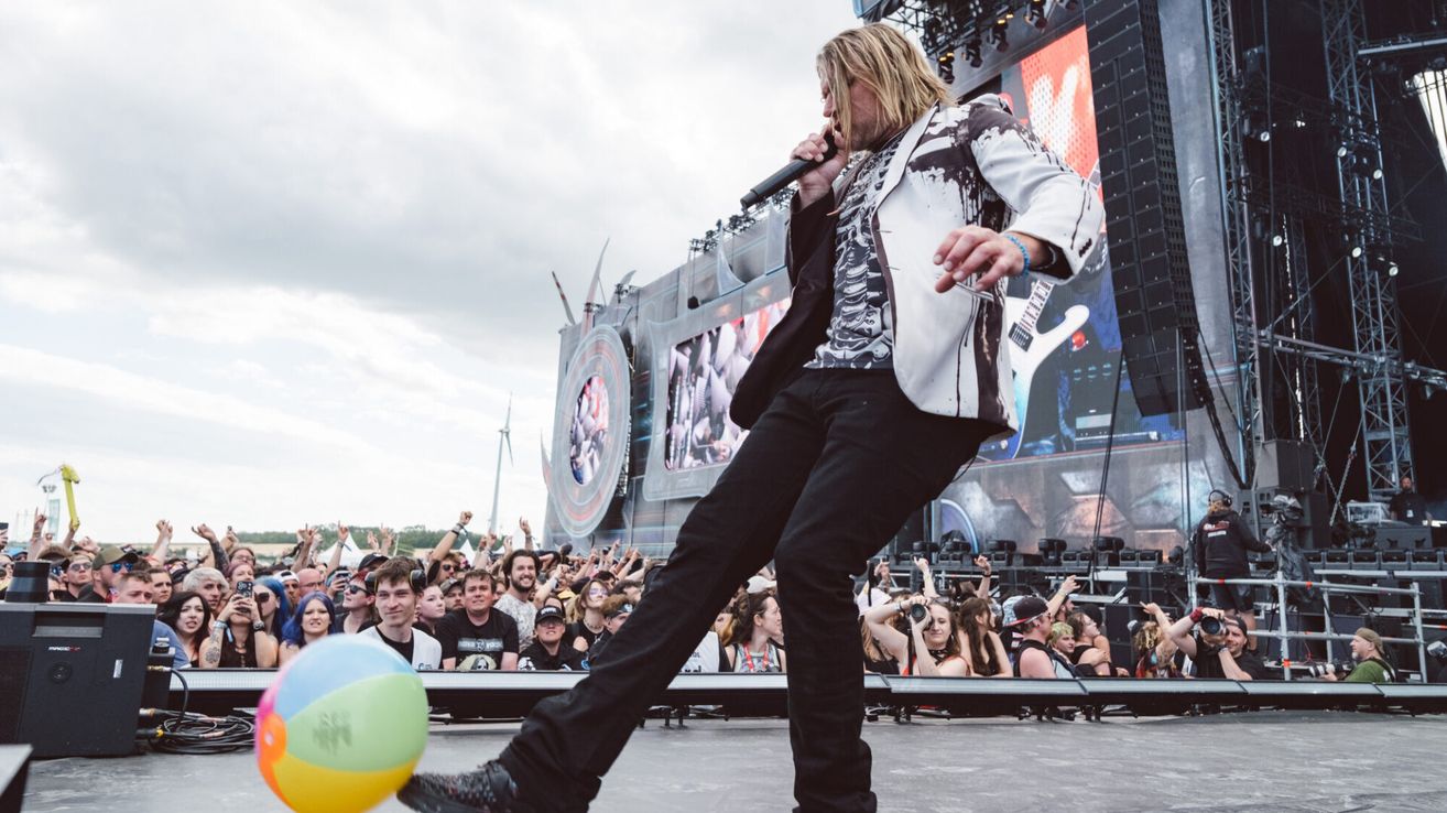 Eine Rockband tritt auf der Bühne auf, während eine große Menge jubelt. Der Leadsänger hält ein Mikrofon und steht mit einem erhobenen Bein. Ein bunter Strandball ist im Vordergrund.