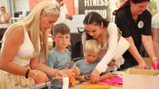 A woman and a girl are teaching two boys to cut paper. The children are sitting at a table with paper and scissors.