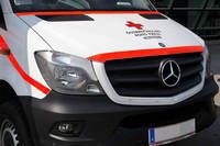 Die Vorderseite eines weißen und orangefarbenen Mercedes-Benz-Transporters mit rotem Kreuzsymbol auf der Motorhaube, Kennzeichen am vorderen Stoßfänger.