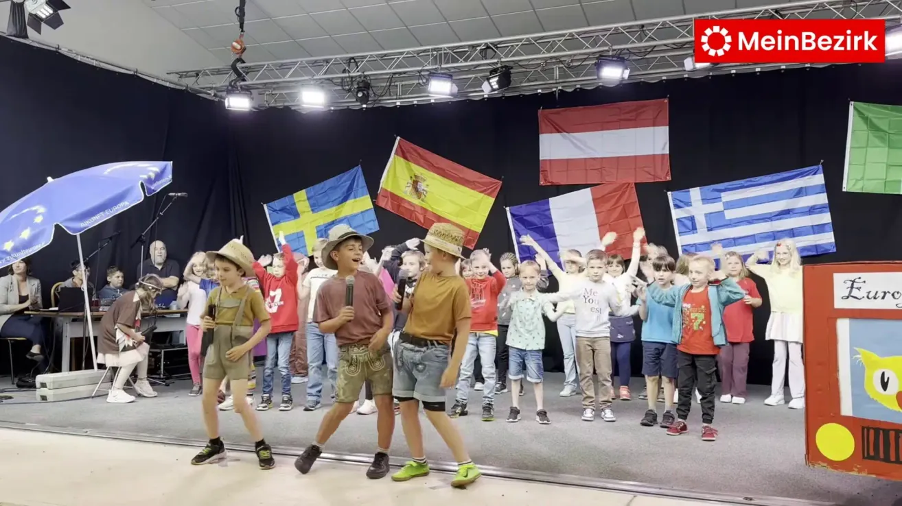 Eine Gruppe von Kindern auf der Bühne mit Flaggen verschiedener Länder, darunter Spanien, Frankreich und Österreich, einige tragen Hüte, halten Flaggen und singen.