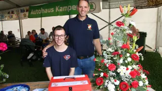 Two men are standing in front of a table with a red donation box and flowers. One man has a smiling face, and the other has glasses and a watch. Behind them, a white tent with banners is visible.