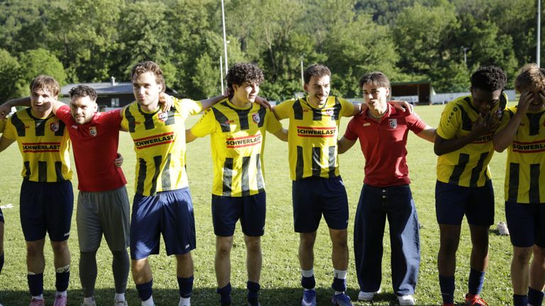 Five young men pose for a photo on a grassy field. Four wear yellow and blue sports uniforms, while the other wears a red shirt. Trees and a building are in the background.
