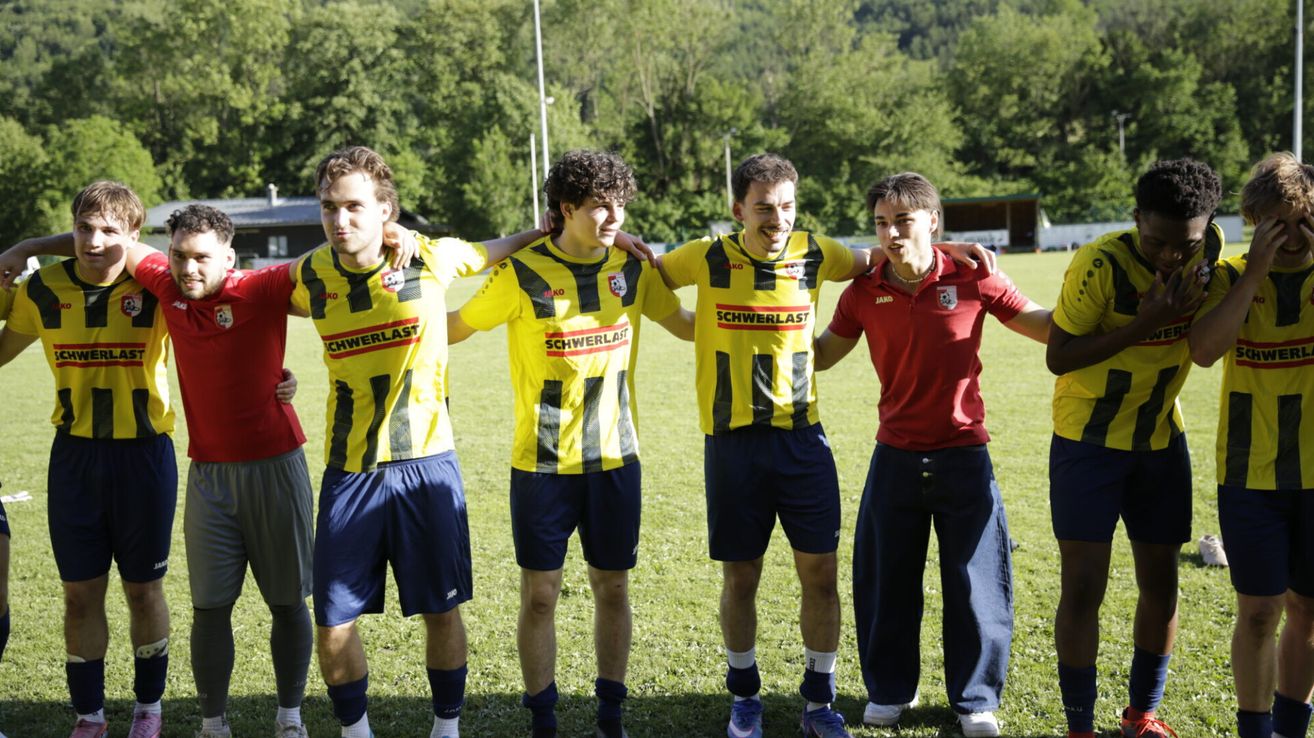 Five young men pose for a photo on a grassy field. Four wear yellow and blue sports uniforms, while the other wears a red shirt. Trees and a building are in the background.