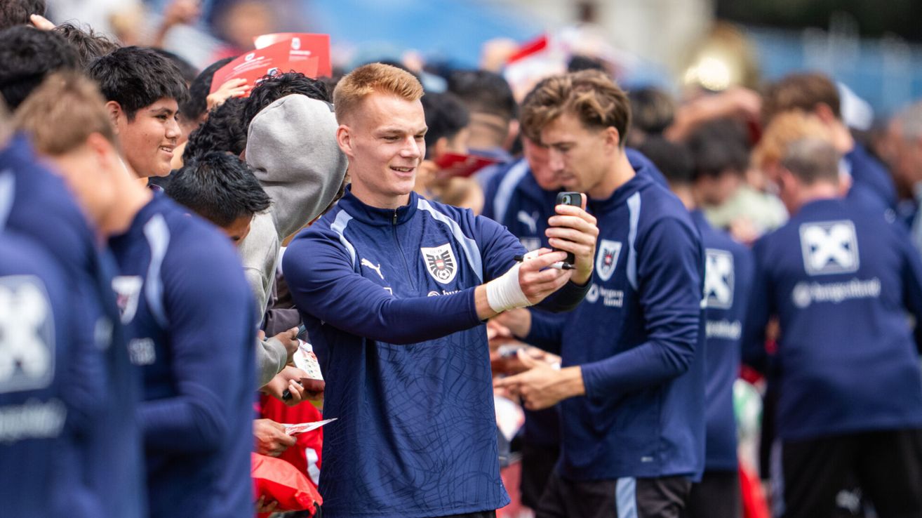 Ein Fußballspieler in Blau macht ein Selfie mit Fans bei einem Spiel. Dahinter warten andere, um ihn zu treffen.