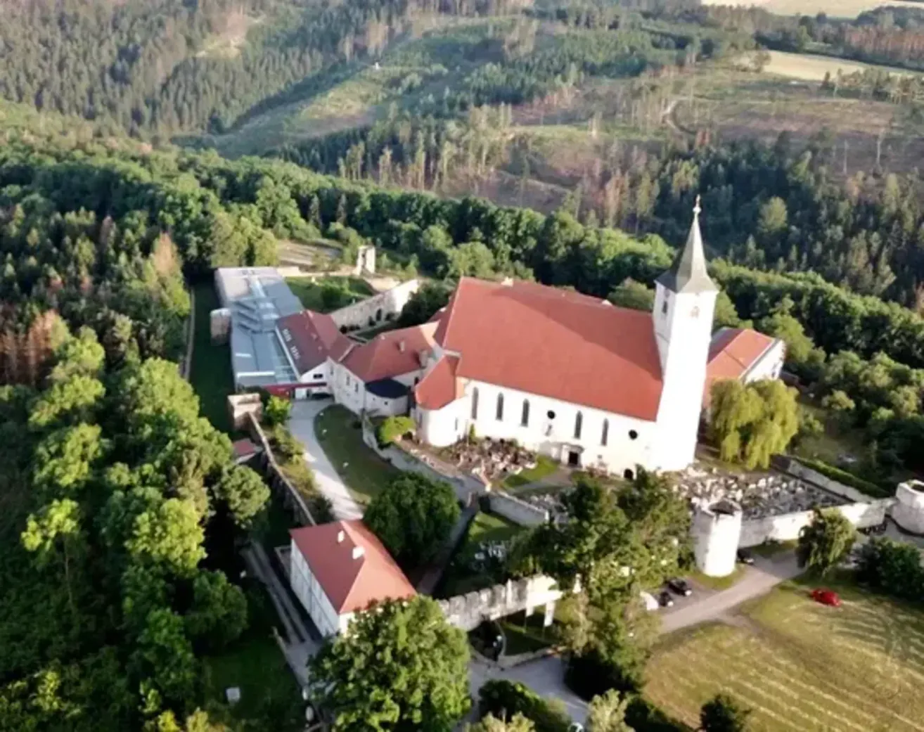 Ein Luftbild einer Kirche mit rotem Dach und Turm, umgeben von grünen Bäumen und Gebäuden in einer ländlichen Gegend.