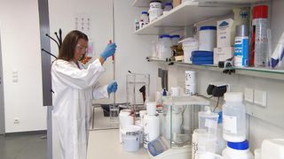 A woman in a lab coat is standing in front of a white countertop, using a pipette to measure liquid. Various lab equipment and chemicals are present.