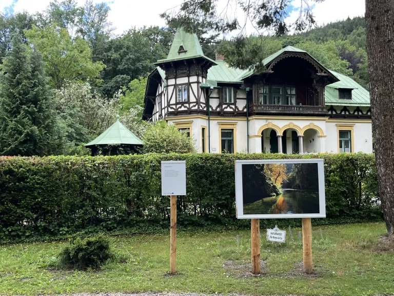 Ein Bilderrahmen steht in einem Garten und zeigt eine Landschaftsansicht. Dahinter befindet sich ein großes Haus mit grünen Dächern, umgeben von Hecken und Bäumen.
