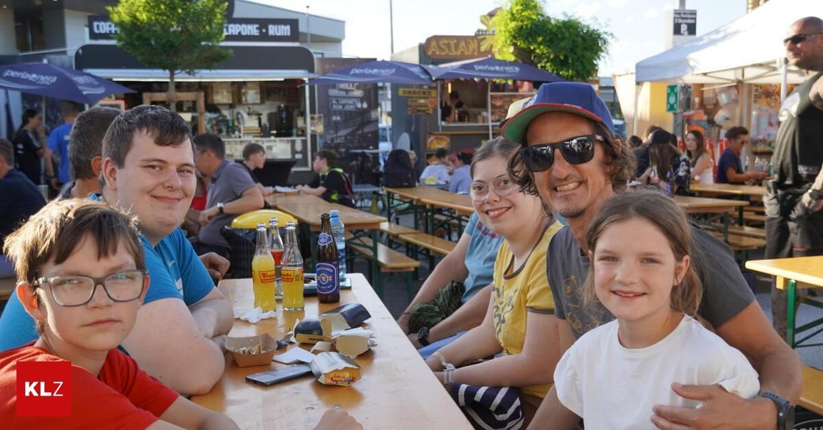 Eine vierköpfige Familie sitzt an einem Tisch im Freien mit Essen und Getränken. Sie lächeln und posieren für ein Foto. Dahinter befinden sich Imbissstände mit blauen Schirmen.