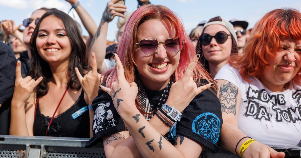 Eine Frau mit pinkem Haar und Tattoos lächelt in die Kamera und macht Friedenszeichen mit den Händen. Sie ist von anderen Konzertbesuchern im Hintergrund umgeben.