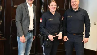 Drei Personen, zwei in Polizeiuniformen und eine in einem grauen Anzug, lächeln und posieren für ein Foto vor einer kunstvollen Holzwand.