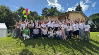 A group of people in white shirts pose for a photo in a grassy field. They are raising their hands. Behind them is a yellow house with a tree. There are balloons and trees in the background.