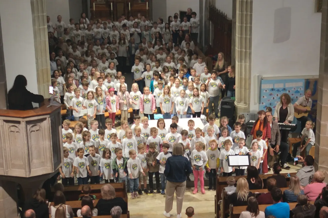 Ein Chor von Kindern in passenden weißen T-Shirts mit Logos steht in einer Kirche, angeführt von einem Erwachsenen mit einem Notenständer. Das Publikum sitzt in Reihen hinter ihnen.