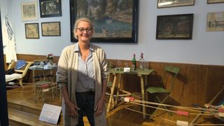 A woman in a blue shirt and glasses stands in a room, smiling. Behind her, a wall displays several paintings, including one of a water scene. A table with wine glasses and a bottle is in front, and two chairs are to the sides.