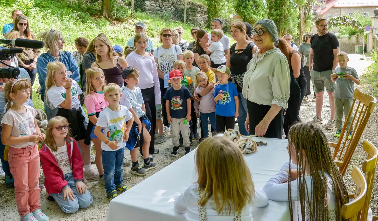 Eine Gruppe von Kindern und Erwachsenen ist um einen Tisch mit einem Fellartikel versammelt, wahrscheinlich für eine Bildungsveranstaltung. Die Frau vorne ist wahrscheinlich die Gastgeberin.