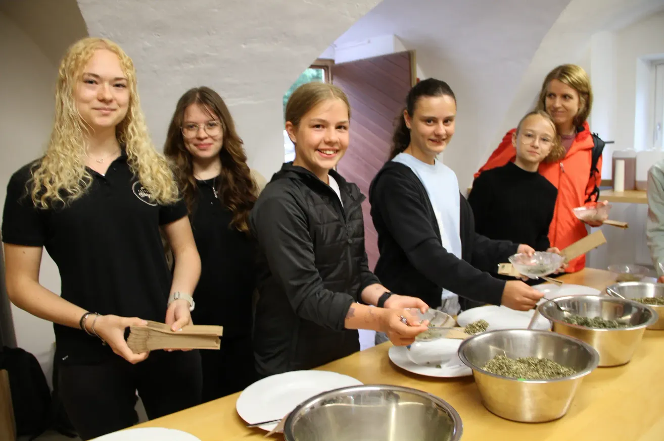 Eine Gruppe junger Frauen bereitet Essen in einem Raum zu. Sie lächeln und scheinen es zu genießen. Schüsseln, Teller und andere Utensilien stehen auf einem Tisch.