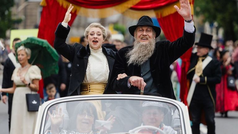 Zwei Personen, gekleidet in altmodischer Kleidung, fahren in einem Cabrio und winken während einer Parade der Menge zu.