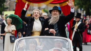 Zwei Personen, gekleidet in altmodischer Kleidung, fahren in einem Cabrio und winken während einer Parade der Menge zu.